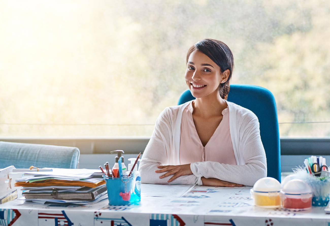 A similing teacher sits at her classroom desk.