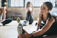 Three women stretching in a gym.  The most prominent lady is smiling while doing a sitting single leg hamstring stretch.